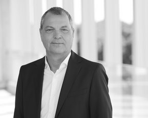 Ein Mann in einem schwarzen Anzug steht vor einer hellen, modernen Architektur. Er hat kurzes, graues Haar und schaut direkt in die Kamera.