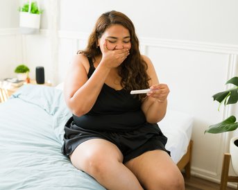 Eine Frau sitzt auf einem Bett in einem hellen Raum und hält einen Schwangerschaftstest in der Hand. Sie hat eine überraschte Miene und trägt einen schwarzen Schlafanzug. Pflanzen und Möbel sind im Hintergrund sichtbar.