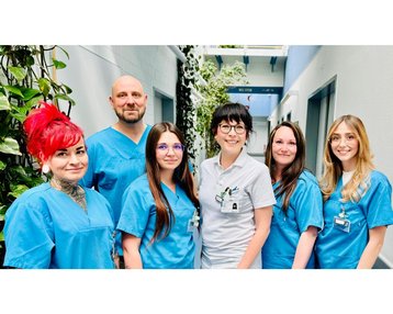 Sechs Personen in blauen Arbeitskleidung stehen in einem Flur mit Pflanzen im Hintergrund. Sie lächeln in die Kamera, während sie sich in einer Gruppe positionieren.