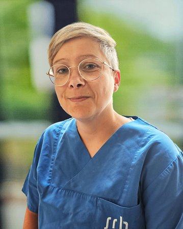 Eine Person in blauer Arbeitskleidung mit Brille sitzt lächelnd. Der Hintergrund ist unscharf, zeigt jedoch eine helle Umgebung.