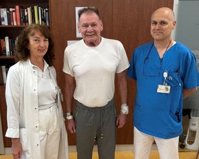 Drei Personen stehen in einem Krankenhauszimmer: eine Frau in weißem Kittel, ein Mann in einem T-Shirt und ein Mann in blauer Kleidung.