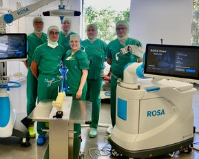 Sechs medizinische Fachkräfte in grüner OP-Kleidung stehen um einen chirurgischen Roboter namens ROSA in einem Operationssaal.