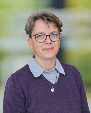 Eine Person mit kurzen grauen Haaren und Brille trägt einen lila Pullover und steht vor einem unscharfen Hintergrund.