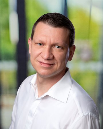 Ein Mann mit kurzen, braunen Haaren und einem weißen Hemd lächelt in die Kamera, vor einem unscharfen, hellen Hintergrund.