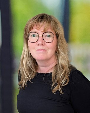 Eine Frau mit langen, blonden Haaren und Brille trägt ein schwarzes Oberteil und einen gestreiften Rock. Sie steht vor einer unscharfen Hintergrundszene.
