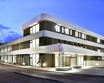 Moderne, mehrstöckige Gebäude mit einer hellen Fassade und großen Fenstern, beleuchtet bei Dämmerung.