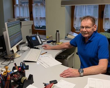 Ein älterer Mann sitzt an einem Schreibtisch mit Computer und Büromaterialien. Er trägt ein blaues Shirt und blickt zur Kamera.