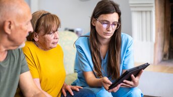 Eine Pflegekraft zeigt älteren Menschen auf einem Tablet Informationen. Die Gruppe sitzt in einem hellen Raum.