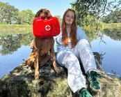 Eine Person - Auszubildende Hedi - sitzt auf einem Stein am Kaimberger Teich, neben ihr ein Hund mit einer roten Erste-Hilfe-Box im Maul.