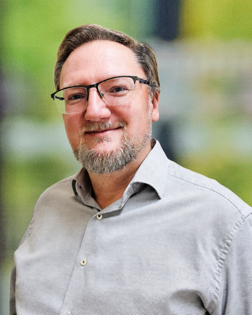 Ein Mann mit Brille und Bart trägt ein helles Hemd und lächelt. Der Hintergrund zeigt unscharfe grüne und graue Töne.