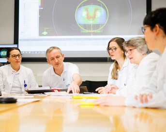Ein Team von Fachleuten in weißen Kitteln diskutiert an einem Tisch. Ein Mann zeigt auf ein digitales Display im Hintergrund, das medizinische Daten anzeigt. Mehrere Personen hören aufmerksam zu und nehmen Notizen.
