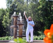 Eine Person - Auszubildende Aylin - steht vor einem Springbrunnen im Dahliengarten Gera und hebt eine Infusionsflasche in die Luft. Umgeben von Bäumen und Blumen.