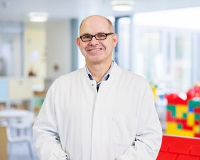 Ein Mann in einem weißen Kittel steht lächelnd in einem Raum mit bunten Spielzeugen und Möbeln, die für Kinder geeignet sind.