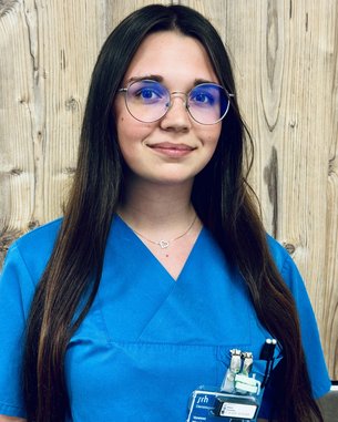 Eine junge Frau mit langen, dunklen Haaren trägt ein hellblaues medizinisches Oberteil und runde Brillen mit blauen Gläsern. Sie hat eine Halskette mit einem Herzanhänger und ein Namensschild an ihrer Kleidung.