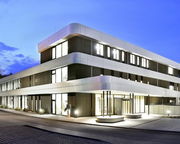 Moderne dreigeschossige Gebäude mit einer eleganten Fassade, beleuchtet bei Dämmerung, umgeben von einer gepflasterten Straße und Bäumen.