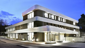 Moderne dreigeschossige Gebäude mit einer eleganten Fassade, beleuchtet bei Dämmerung, umgeben von einer gepflasterten Straße und Bäumen.