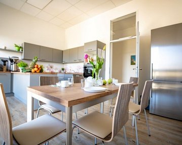 Moderne Küche mit grauen Schränken, einem Holztisch und Stühlen. Auf dem Tisch stehen Geschirr und frische Blumen. Im Hintergrund sind Obst und Küchengeräte sichtbar.