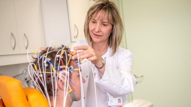 Eine medizinische Fachkraft befestigt Elektroden an der Kopfhaut eines Patienten, der in einem Behandlungsraum sitzt. Die Elektroden sind mit bunten Drähten verbunden und dienen wahrscheinlich zur Durchführung eines EEGs oder ähnlicher Tests.
