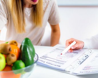 Eine Person zeigt auf ein Dokument in einem Ordner, während eine andere Person aufmerksam zuhört. Eine Schüssel mit Obst steht auf dem Tisch.