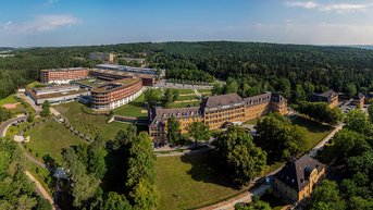 Luftaufnahme eines großen Gebäudekomplexes umgeben von Bäumen und Wiesen, mit modernen und historischen Architekturelementen.