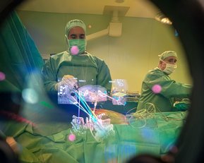 Ein Chirurg in OP-Kleidung arbeitet an einem Patienten, sichtbar durch ein chirurgisches Instrument. Ein zweiter Chirurg ist ebenfalls im Hintergrund zu sehen.
