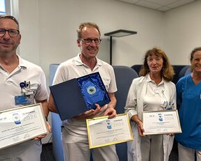 Vier Personen in Arbeitskleidung halten Auszeichnungen und Urkunden in einem Büro. Eine Person zeigt eine Plakette.