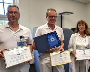 Vier Personen in Arbeitskleidung halten Auszeichnungen und Urkunden in einem Büro. Eine Person zeigt eine Plakette.