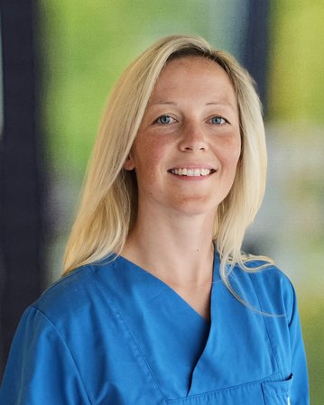 Eine Person mit blonden Haaren trägt ein blaues medizinisches Outfit und lächelt freundlich in einem hellen Raum.