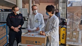 Drei Personen in Schutzmasken stehen um einen Tisch mit Medikamenten, während sie Dokumente besprechen. Im Hintergrund sind Kartons sichtbar.