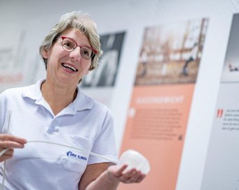 Chefärztin Prof. Stroh trägt ein weißes Polohemd und hält ein medizinisches Gerät - einen Magenballon - in der Hand. Im Hintergrund sind Informationswände mit Texten und Bildern zu sehen.