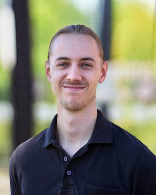 Junger Mann mit langen Haaren, der in einem schwarzen Poloshirt lächelt, vor einer unscharfen grünen Hintergrundlandschaft.