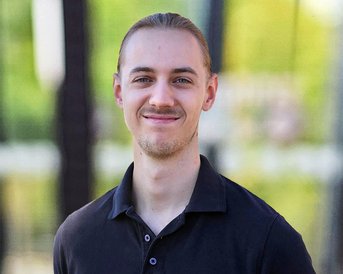 Junger Mann mit langen Haaren, der in einem schwarzen Poloshirt lächelt, vor einer unscharfen grünen Hintergrundlandschaft.