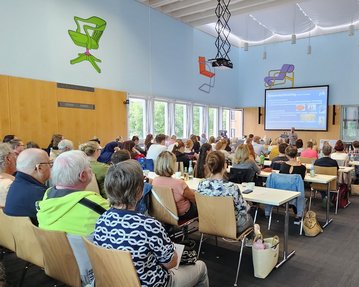 Eine große Gruppe von Menschen sitzt in einem hellen Raum und hört einer Präsentation auf einem Bildschirm zu.