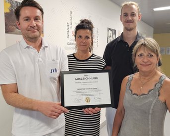 Vier Personen halten eine Urkunde mit der Aufschrift "AUSZEICHNUNG". Sie stehen in einem Innenraum, wobei zwei Personen in weißen Arbeitskleidung und zwei in Alltagskleidung gekleidet sind.