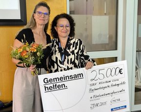 Zwei Frauen stehen zusammen, eine hält einen Blumenstrauß, die andere präsentiert einen Scheck über 25.000 Euro für eine Einrichtung.