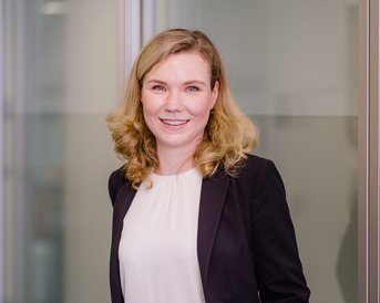 Eine Frau mit blonden, lockigen Haaren steht in einem modernen Büro. Sie trägt einen schwarzen Blazer über einer weißen Bluse und hat die Hände in den Taschen ihrer schwarzen Hose.