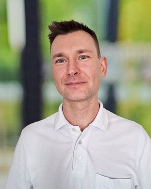 Ein Mann mit kurzem Haar trägt ein weißes Polohemd mit einer Brusttasche. Er lächelt und steht vor einem unscharfen, hellen Hintergrund.