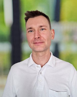 Ein Mann mit kurzem Haar trägt ein weißes Polohemd mit einer Brusttasche. Er lächelt und steht vor einem unscharfen, hellen Hintergrund.