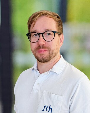 Mann mit Brille und kurzen Haaren trägt ein weißes Polohemd mit dem Logo "srh". Hintergrund unscharf, grüne Töne sichtbar.
