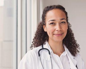 Eine lächelnde Ärztin mit lockigem Haar trägt einen weißen Kittel und ein Stethoskop um den Hals. Sie steht vor einem Fenster.