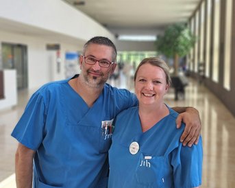 Zwei medizinische Fachkräfte in blauen Kitteln stehen lächelnd in einem Krankenhausflur.