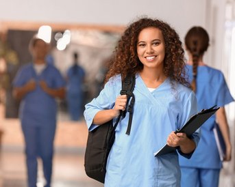 Eine junge Frau in blauer Arbeitskleidung hält eine Mappe in der Hand und lächelt, während sie in einem Flur mit anderen Personen steht.
