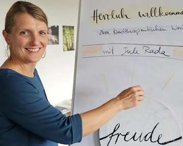 Eine lächelnde Frau zeigt auf ein Flipchart mit der Aufschrift „Herzlich willkommen zum künstlerischen Workshop mit Jule Rode“ und dem Wort „freude“.