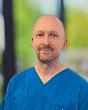Ein Mann in einem blauen medizinischen Outfit steht lächelnd vor einem unscharfen Hintergrund. Er hat kurze Haare und einen gepflegten Bart.