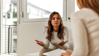 Eine Frau sitzt auf einem Sofa und spricht mit einer anderen Person, die ihr gegenüber sitzt. Sie wirkt nachdenklich und gestikuliert.