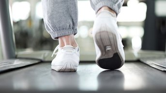 Eine Person in grauen Jogginghosen läuft auf einem Laufband, während sie weiße Sportschuhe trägt. Der Fokus liegt auf den Füßen und dem Laufband.