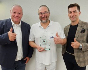 Drei Männer stehen zusammen und zeigen Daumen hoch. Der mittlere Mann hält einen Preis in der Hand. Alle lächeln und tragen formelle Kleidung.
