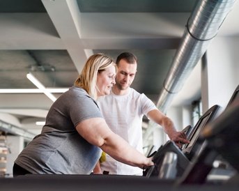 Eine Frau mit Übergewicht beginnt das Training auf dem Laufband, ein Trainer gibt Hinweise.