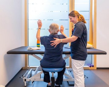 Eine Physiotherapeutin hilft einem älteren Patienten, der auf einem Behandlungstisch sitzt und seine Arme hebt. Der Raum ist hell und modern, mit großen Fenstern und minimalistischem Design.