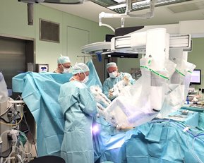 Ein chirurgisches Team in einem Operationssaal führt einen Eingriff durch. Die Ärzte tragen Schutzkleidung und Masken, während sie sich auf den Patienten konzentrieren, der von blauen Tüchern umgeben ist.